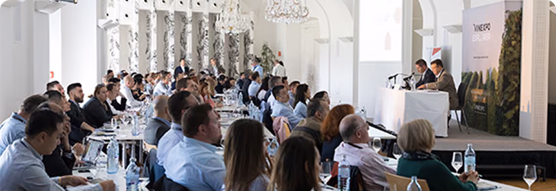 Audience seated at tables attentively watching two speakers presenting on a stage in a bright conference room with white walls and chandeliers.