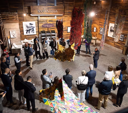 Group of people standing in a circular formation inside a wooden room, observing colorful large hanging art installations resembling abstract animal shapes, with framed artwork on the walls and a sign reading 'Santa Julia'.