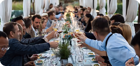 People sitting at a long outdoor dining table under a canopy, raising their glasses for a toast in a festive gathering.