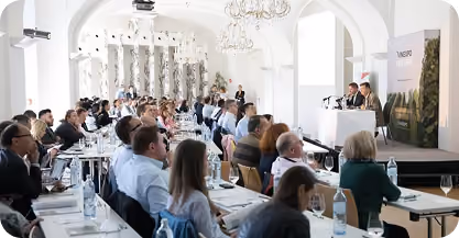 Audience seated at long tables in a bright, elegant room with arched ceilings and chandeliers, watching a panel discussion with two speakers at a podium.