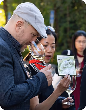 A man wearing a gray flat cap and dark jacket smelling white wine in a glass while a woman beside him holds a wine bottle label and a glass of wine, with other people blurred in the background outdoors.