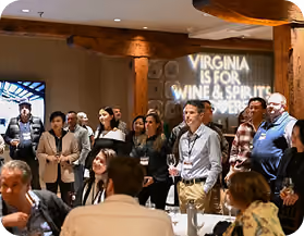 Group of people standing and sitting in a dimly lit room with a sign on the wall that reads 'Virginia is for Wine & Spirits'.