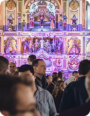 Crowd of people standing in front of a large, colorful, ornate mechanical musical display with the name 'Gaudin & Cie' and 'Paris' visible, decorated with figures and intricate designs.