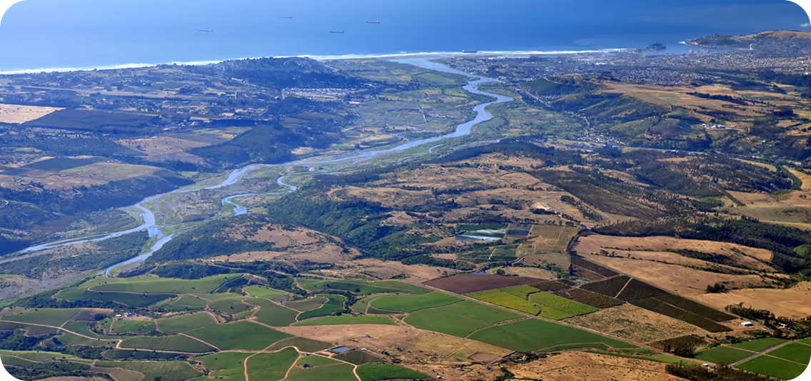 Aerial view of a winding river flowing through a valley with green and brown fields, leading to a coastal city by a calm blue sea with several ships anchored offshore.