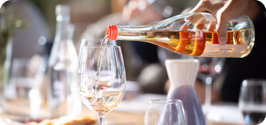 A hand pouring white wine from a bottle with a red cap into a wine glass on a table with blurred background objects and people.