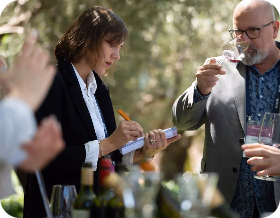 Woman in a black blazer noting something in a notebook with an orange pen while a bearded man in glasses sniffs a glass of red wine, outdoors with blurred green foliage background.