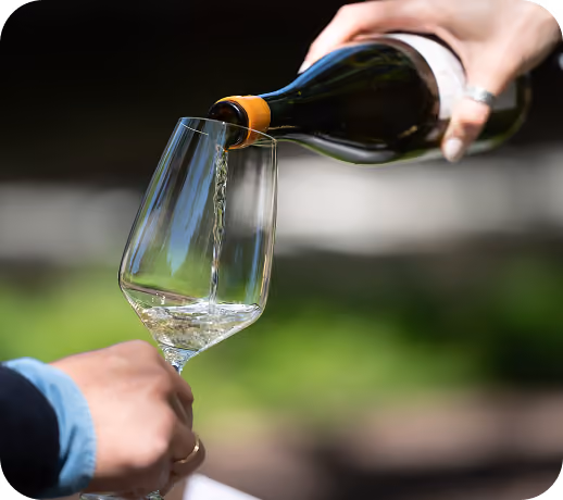A close-up of a hand pouring white wine from a dark green bottle into a tilted wine glass held by another person, with a blurred green and brown background.