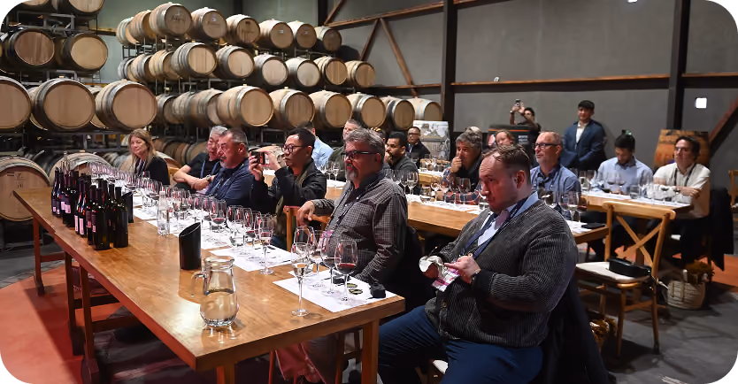 Group of people sitting at wooden tables in a wine cellar with stacked barrels, participating in a wine tasting event with wine bottles and glasses in front of them.
