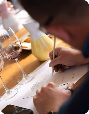 Person writing or filling out a form on a clipboard with a pencil, with two glasses of red wine and a wine aerator on a wooden table nearby.