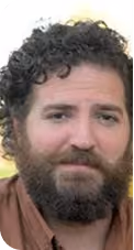 Man with curly dark hair, full dark beard, and neutral expression wearing a brown shirt.
