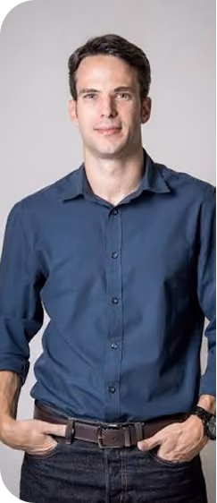 Portrait of a man with short dark hair wearing a dark blue button-up shirt with sleeves rolled up and dark jeans, standing with hands in pockets against a plain light gray background.