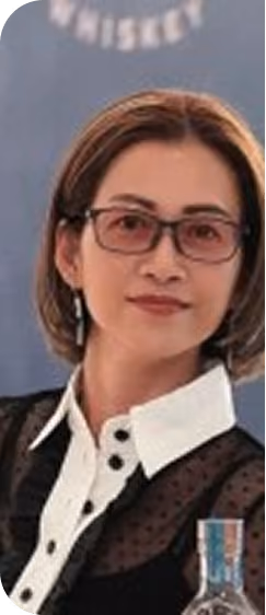 Portrait of a woman with short brown hair wearing glasses, a black top with a white collar and buttons, standing in front of a blurred blue background with partially visible text.