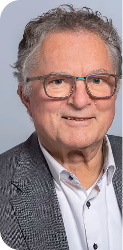 Portrait of an older man with gray curly hair and glasses, wearing a white shirt and gray blazer, smiling slightly against a plain light blue background.