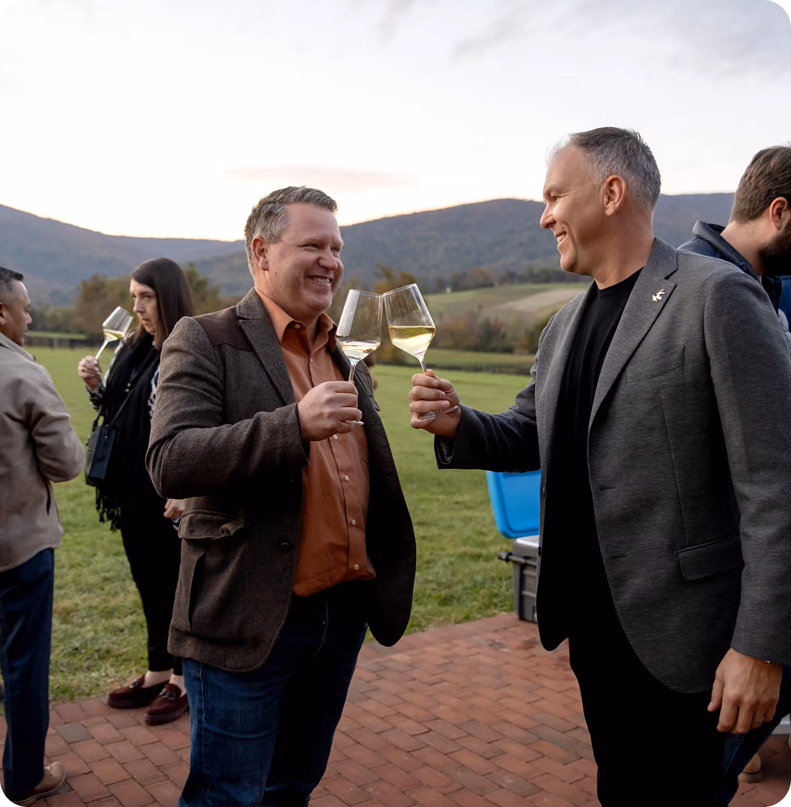 Two men smiling and clinking white wine glasses outdoors on a brick patio with a grassy field and hills in the background during early evening.