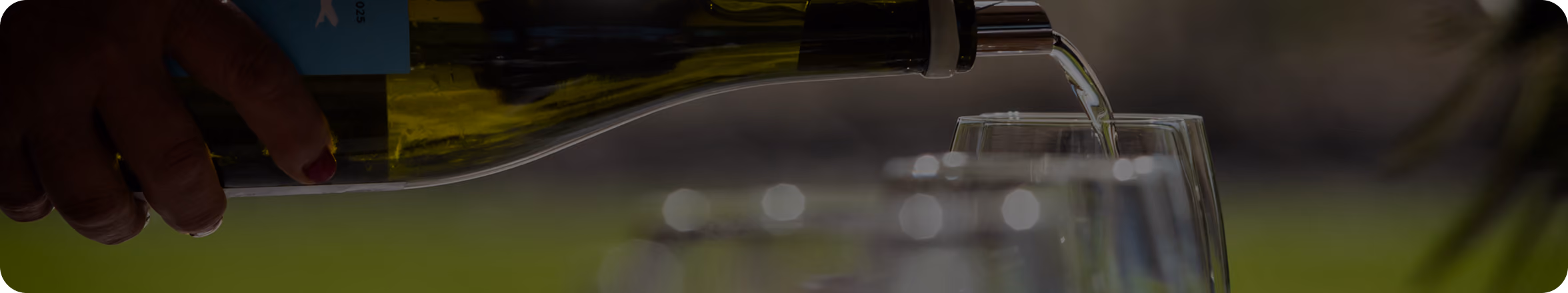Hand pouring white wine from a bottle into a glass outdoors with blurred greenery background.