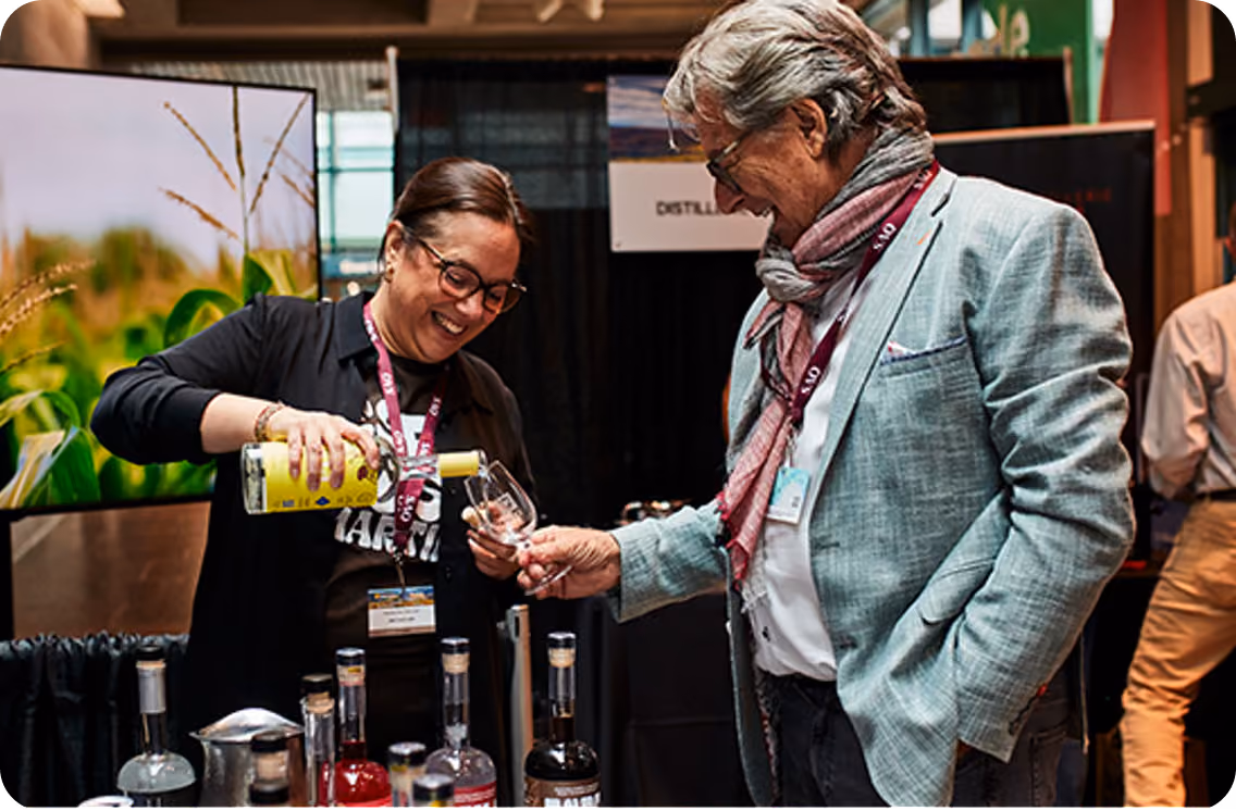 A smiling woman pours a yellow bottled drink into a glass held by a man wearing a light gray suit and scarf at a booth displaying multiple liquor bottles.