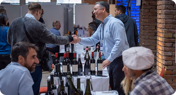 Two men shaking hands across a table with numerous wine bottles displayed, at a wine tasting or event in an indoor venue with brick walls.