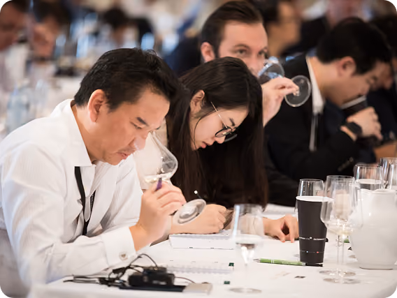 Four people seated at a table, each smelling wine in glasses as part of a wine tasting event, with notebooks and pens in front of them.