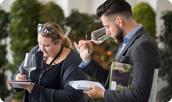 Man in a suit smelling wine in a glass while holding a notebook and a book, with a woman beside him writing in a notebook and holding a wine glass, both in a setting with blurred green plants in the background.