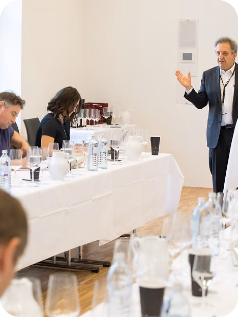 A man in a suit is standing and speaking to seated people at a long table covered with a white tablecloth, with glasses and bottled water on it, in a bright conference room.