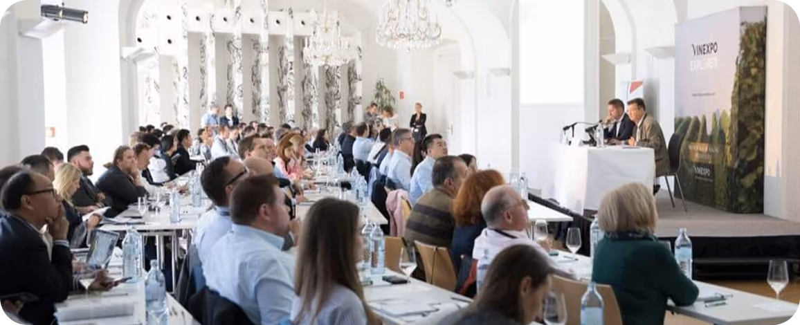 Audience seated at long tables in a bright conference room with chandeliers, listening to two speakers at a podium on stage during a formal presentation.