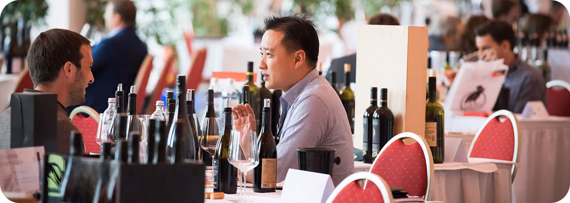 Two men seated at a table with multiple bottles of wine and wine glasses, engaged in conversation at a wine tasting event.