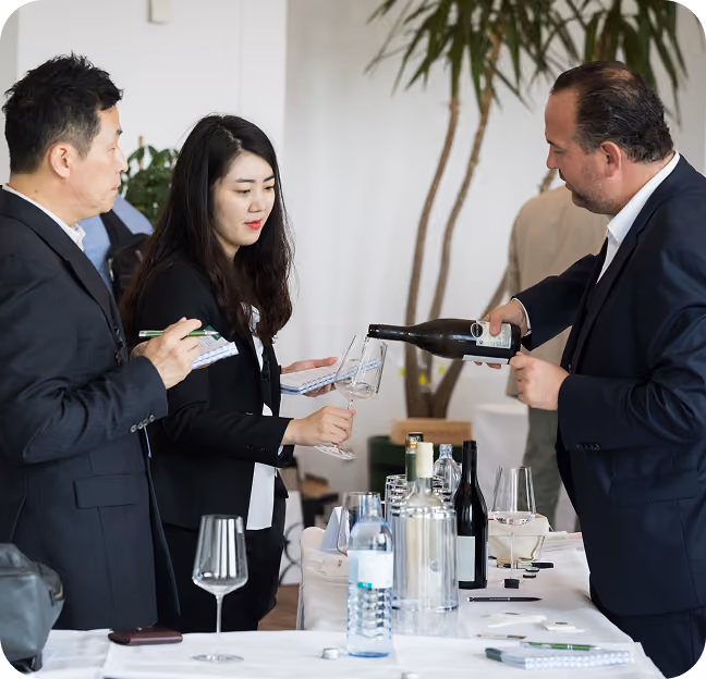Three people at a wine tasting event, with a man pouring wine into a glass held by a woman who takes notes, and another man also observing and writing on a notepad, with bottles and glasses on a table in front of them.