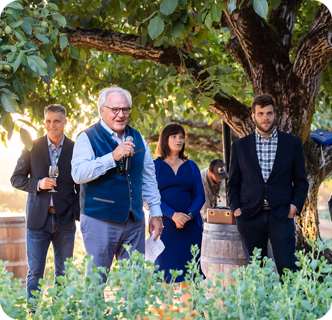 Four people stand outdoors under a leafy tree at sunset during a gathering, with one man in a blue vest speaking into a microphone while others listen.