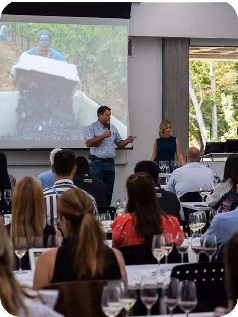 A man with a microphone giving a presentation to an audience seated at tables with wine glasses; a woman stands nearby and a projection screen shows a person pouring grapes into a container outdoors.