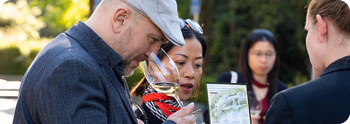 A man wearing a gray cap smelling a glass of white wine while a woman beside him looks at a vineyard map during a wine tasting outdoors, with two other people in the background.