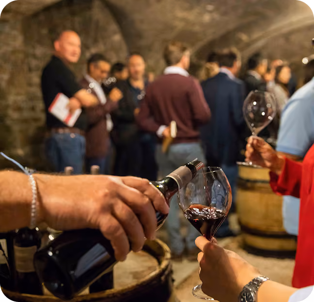 A person pouring red wine into a glass held by another person in a lively wine tasting event with several people conversing in the background inside a rustic cellar.