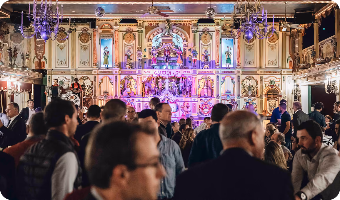 Crowd of people gathered inside an ornate room with a large, brightly lit decorative mechanical orchestra or organ display labeled 'Gaudin & Cie' in the background.