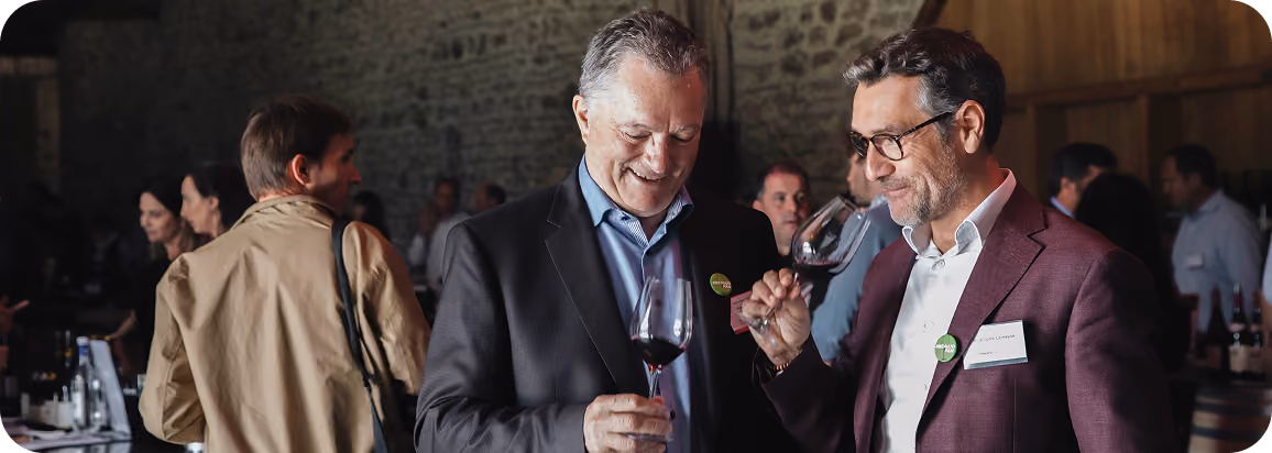 Two middle-aged men in suits holding and smelling glasses of red wine at a social event in a rustic indoor setting with stone walls and other attendees mingling in the background.