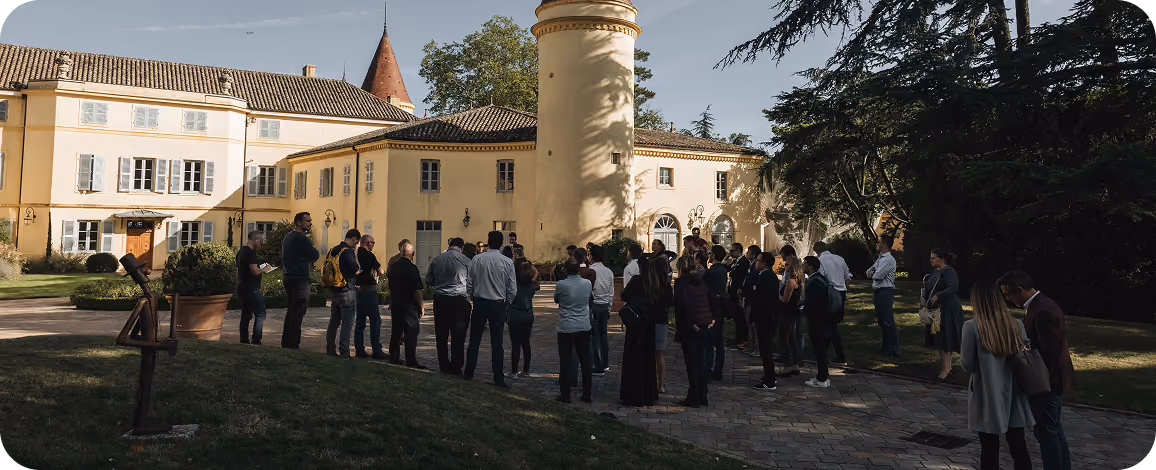 Group of people gathered outdoors in front of a large yellow building with a round tower and tiled roofs during daytime.