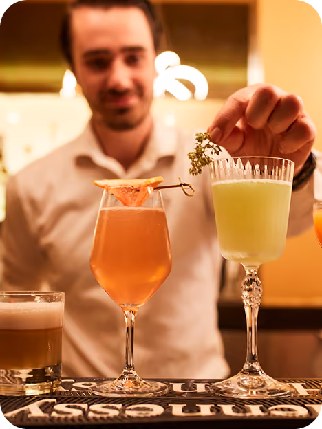 Bartender in a white shirt garnishing a green cocktail with a small sprig of herbs on a bar counter with three different cocktails in front of him.