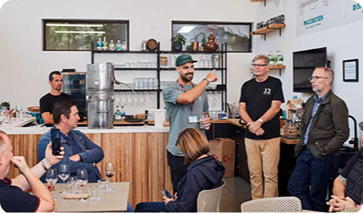 A group of people inside a modern café or tasting room with wooden paneling and shelves with glasses and bottles, one man with a beard and cap is speaking and gesturing while others listen attentively.