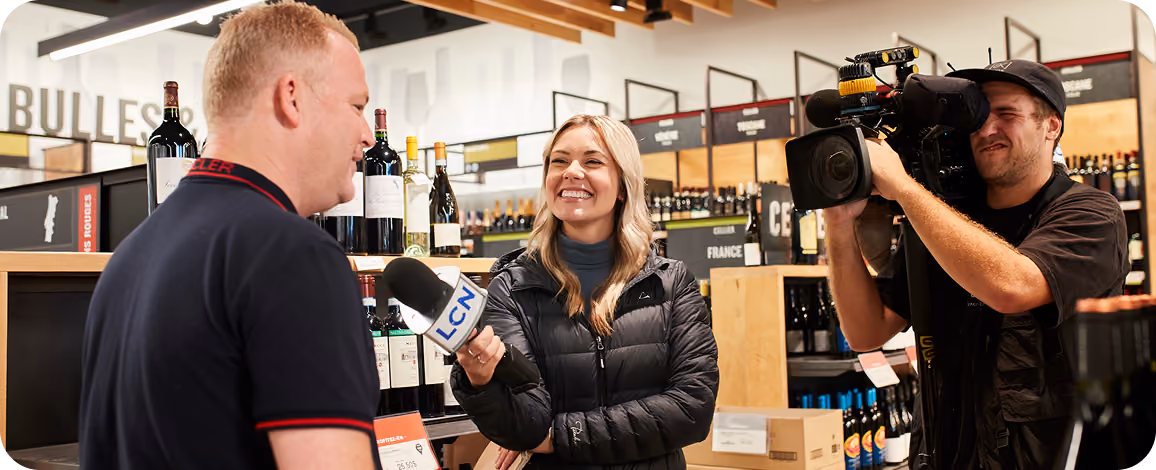 A female reporter holding an LCN microphone interviews a man in a store aisle with wine bottles, while a cameraman films them.