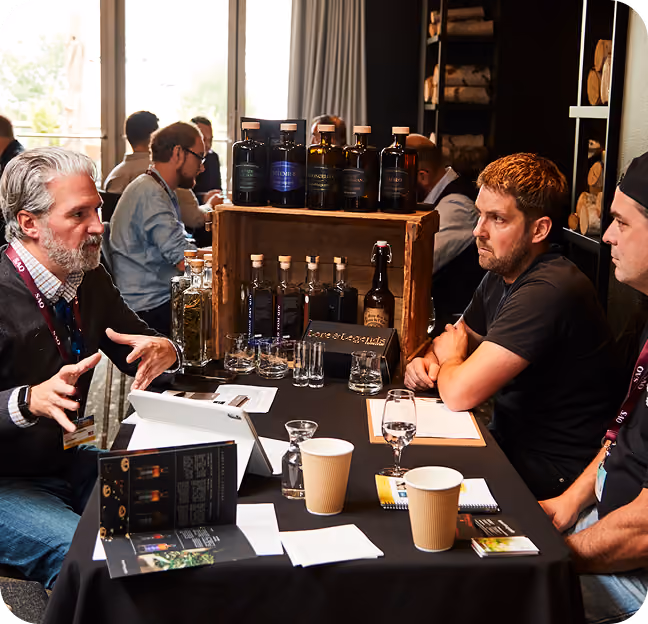 Three men sit around a table in discussion at a trade event or meeting, with bottles of liquor or spirits displayed on a wooden crate at the center, alongside glasses and informational materials.