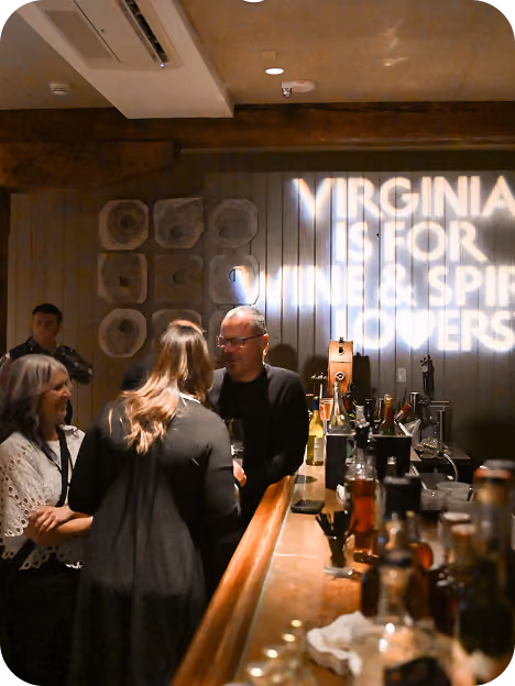 People socializing at a bar with a wooden countertop and bottles of liquor, with a light projection on the wall saying 'VIRGINIA IS FOR WINE & SPIRITS LOVERS.'