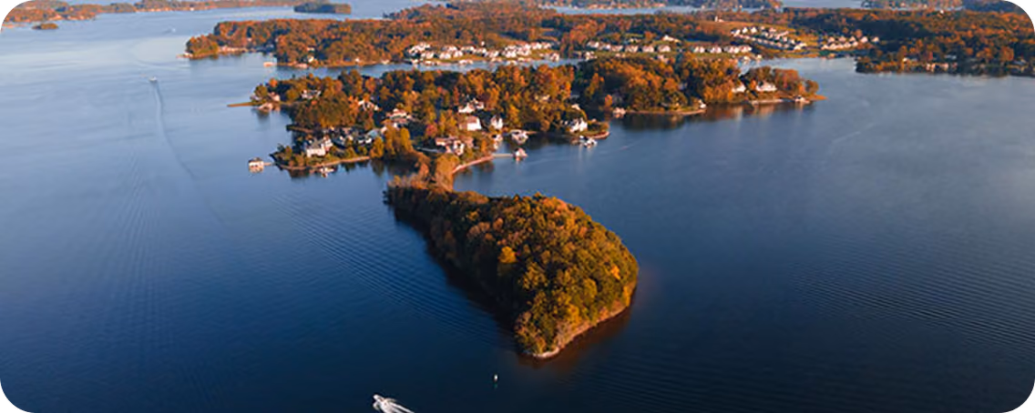Aerial view of a small forested peninsula surrounded by calm water, with a white boat leaving a wake near the shore and houses along the coastline in the background during autumn.