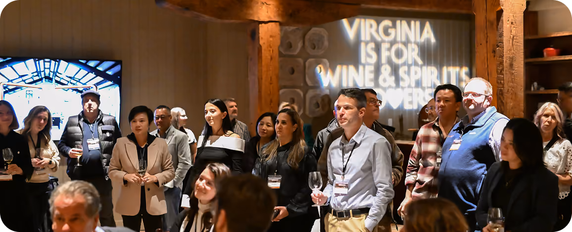 Group of diverse people standing indoors at an event, some holding wine glasses, with a sign on a wall reading 'Virginia is for Wine & Spirits Lovers.'
