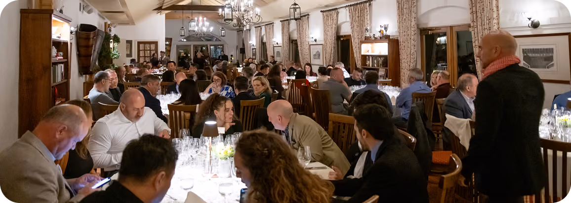 A crowded dining room with many people seated at long tables, engaged in conversation and dining under chandeliers and lantern-style ceiling lights in a warmly lit, elegant setting.