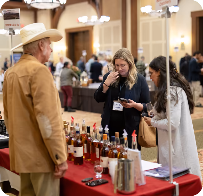 A man in a tan jacket and white cowboy hat stands opposite two women at a table with numerous bottles of liquor, one woman smelling a sample while the other looks on holding a glass, with a sign labeled 'Catoctin Creek' above the table in a large room with other people and tables in the background.