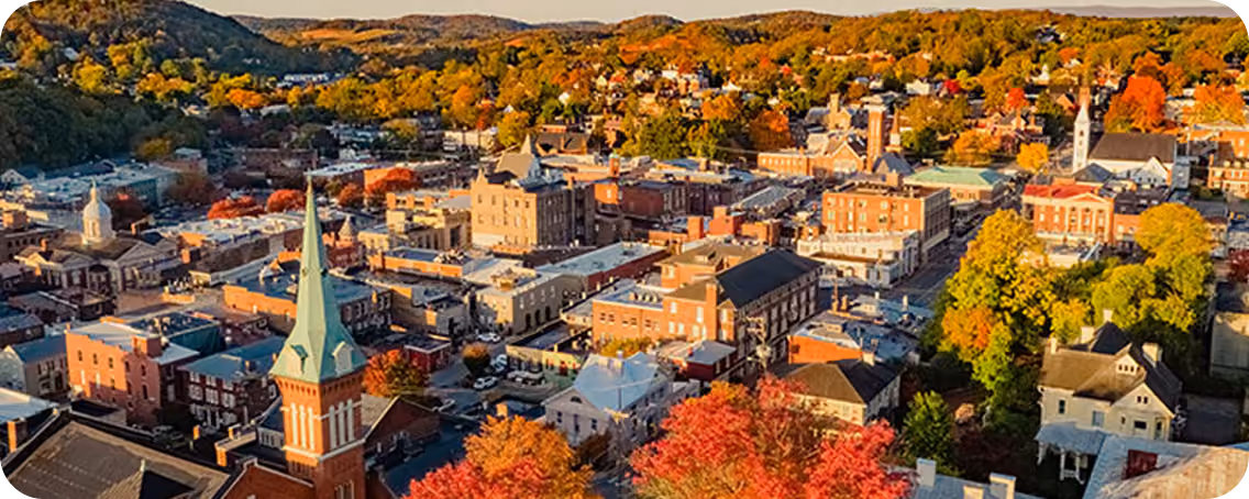 Aerial view of a small town in autumn with colorful fall foliage, historic brick buildings, church steeples, and surrounding hills in the background.