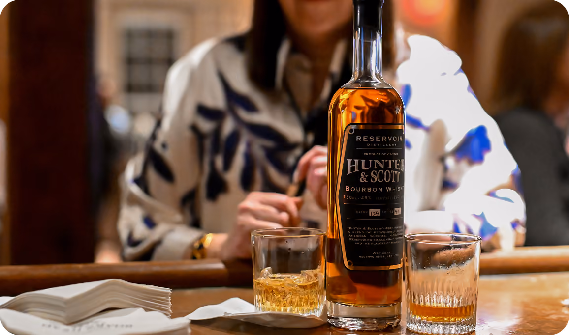 A bottle of Hunter & Scott bourbon whiskey on a bar counter with two whiskey glasses, one partially filled, in front of a person wearing a white and blue patterned jacket.