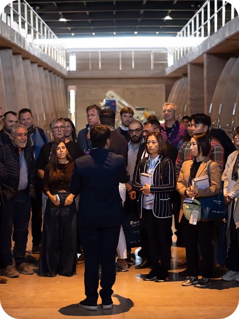 Group of diverse adults attentively listening to a speaker in a spacious industrial-style room with high ceilings and wooden beams.