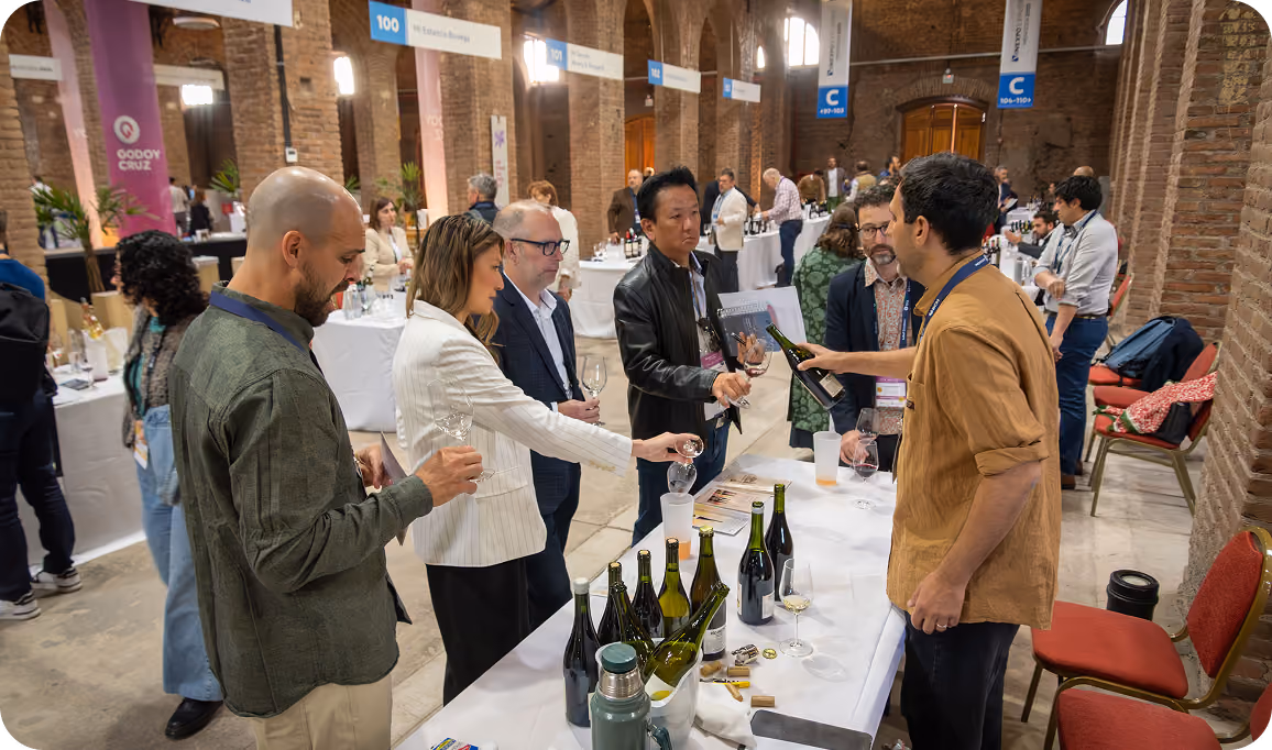 A group of people standing around a table at a wine tasting event in a spacious hall with brick arches, sampling and pouring wine from bottles displayed on the table.