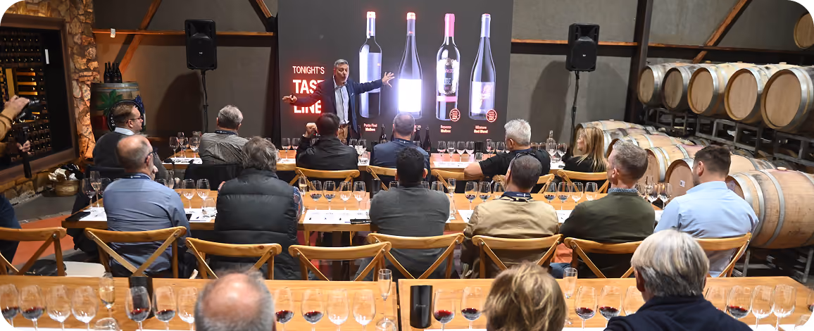 A group of people seated at wooden tables with rows of wine glasses in front of them, watching a presenter standing by a screen displaying enlarged images of four wine bottles during a wine tasting event in a cellar with oak barrels.