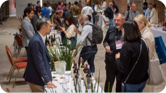 People attending a wine tasting event, standing around tables with bottles of wine and wine glasses in a large indoor venue.
