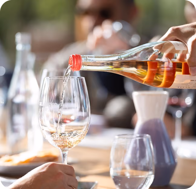 A person pours white wine from a bottle with an orange cap into a wine glass held by another hand, with a blurred background of other glasses and people at a table.